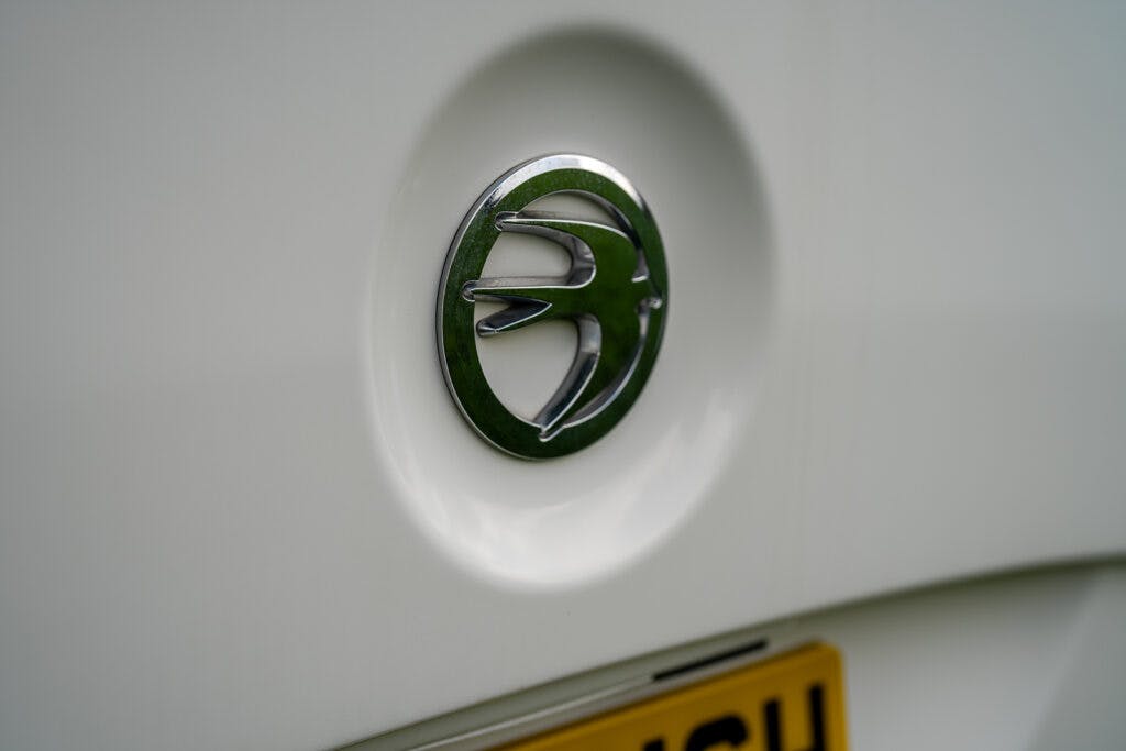 Close-up of a silver car emblem with a design resembling a bird in flight on a 2017 Swift Escape 664. The background shows part of a yellow license plate blurred out and partial white car body surface.