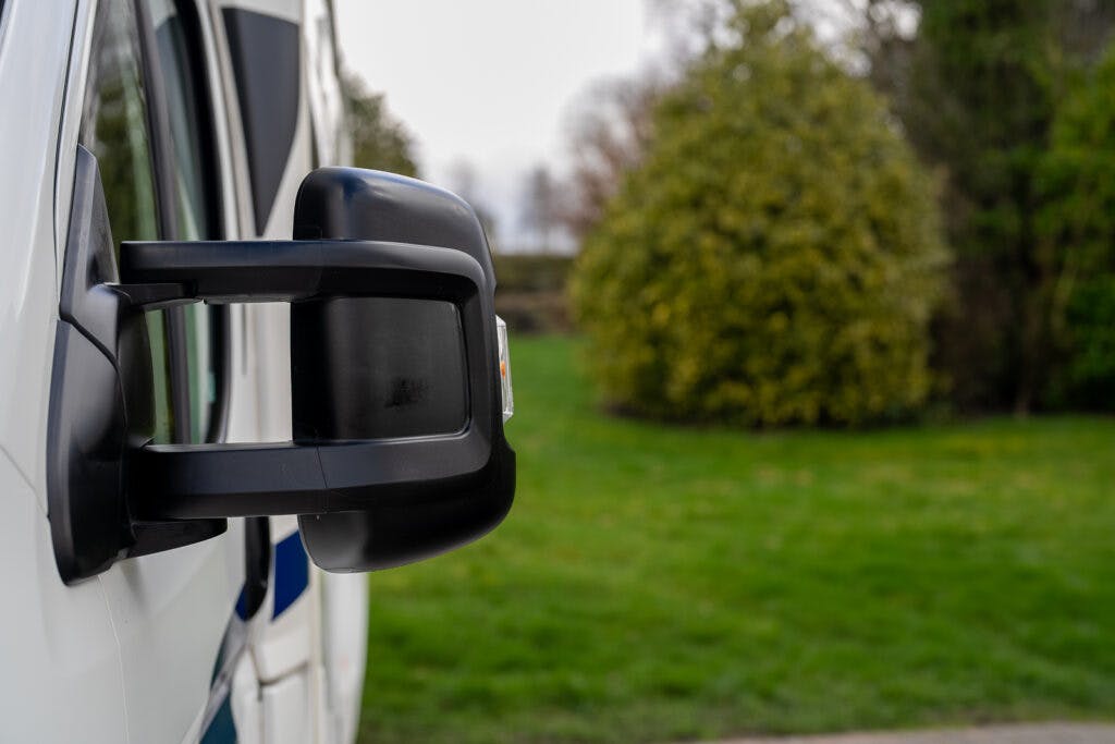 A close-up shot of a black side mirror on a 2017 Swift Escape 664. The background features a grassy area with trees and shrubs on a slightly overcast day. The focus is primarily on the side mirror with the greenery softly blurred in the background.