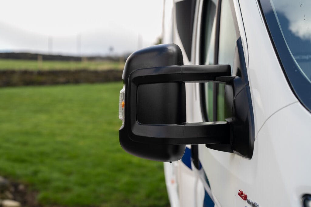 Close-up image of a side mirror on a white 2017 Swift Escape 664. The mirror features a sleek black housing and is seen against a blurred background of a grassy field and overcast sky.
