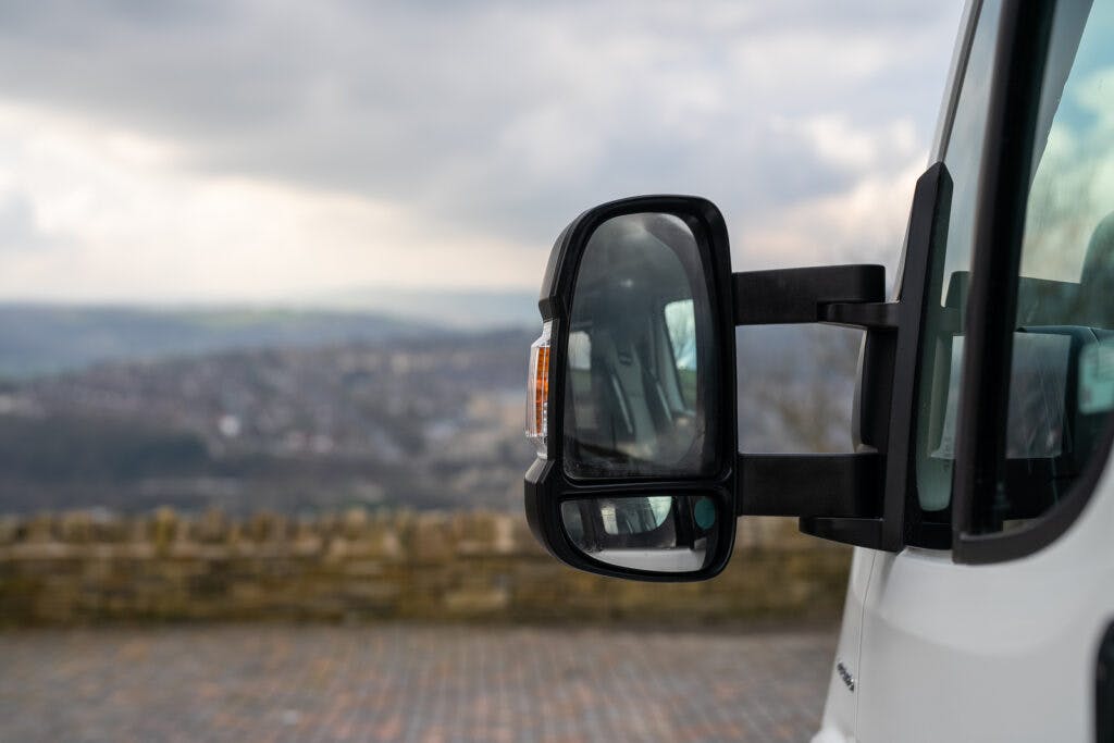 A close-up view of the side mirrors of a white 2017 Swift Escape 664, with a landscape of rolling hills and a cloudy sky in the background. The vehicle is parked on a paved surface with a stone barrier in the distance.