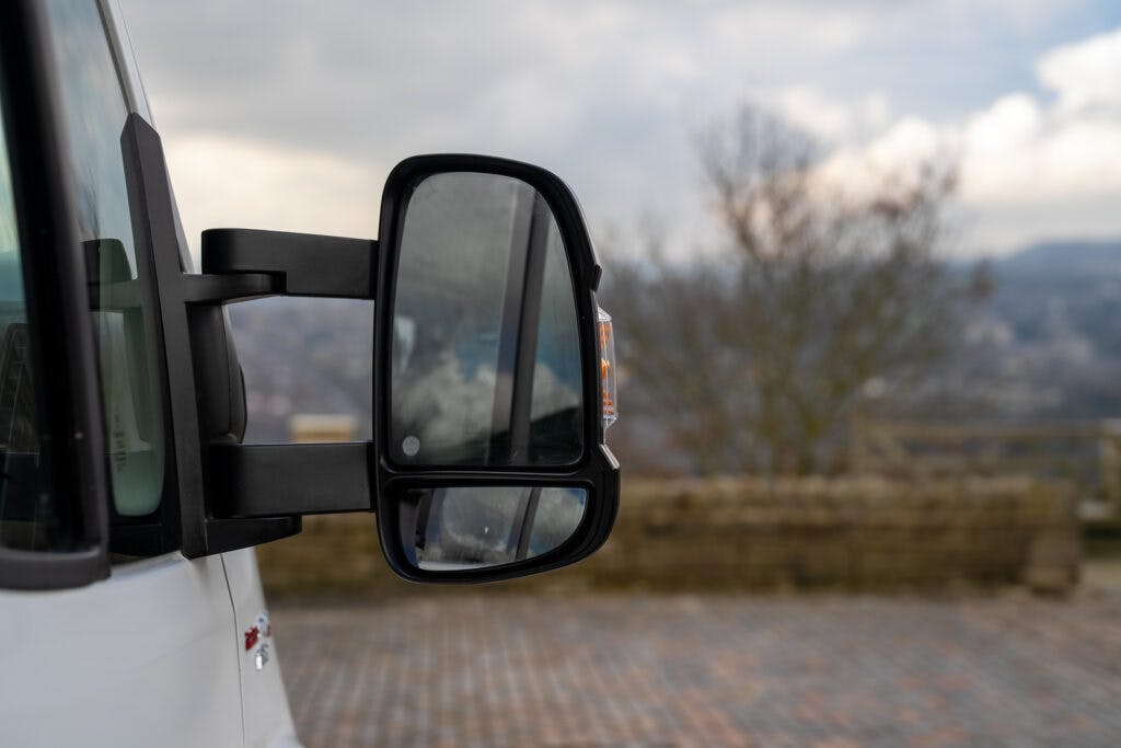 A close-up of the side mirror on a white 2017 Swift Escape 664, showing reflections of the cloudy sky and surrounding scenery. The background is slightly blurred, featuring a stone wall and an out-of-focus landscape. The paved ground is partially visible.