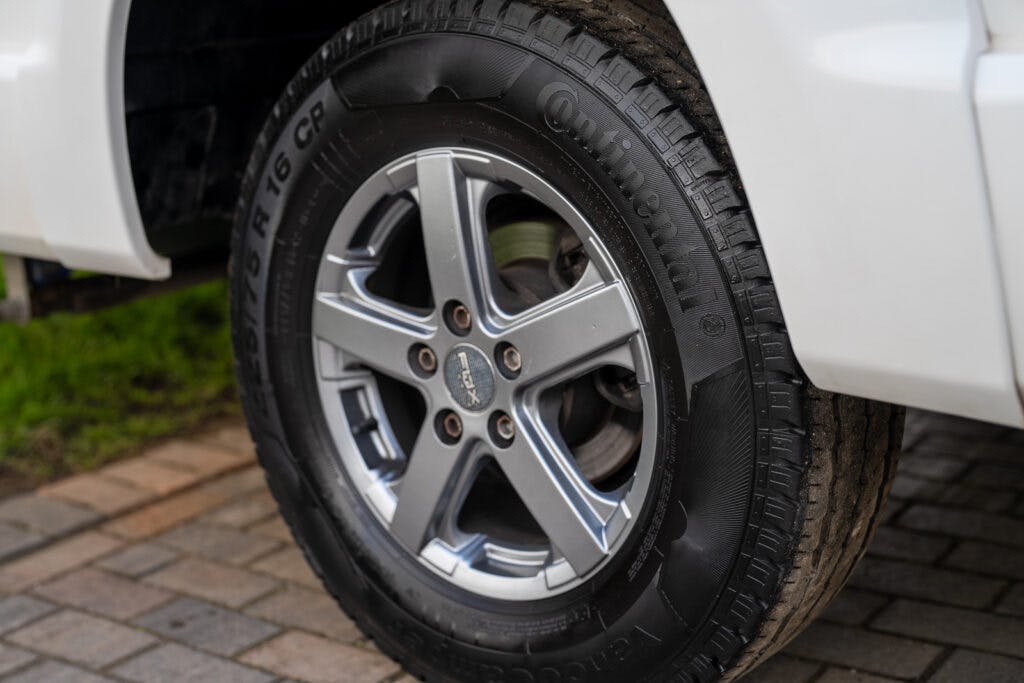 Close-up of a car tire and wheel on a paved surface. The tire is a Continental brand, and the rim has a five-spoke design. The car's white body, likely from the 2017 Swift Escape 664, is partially visible in the background. Some grass can be seen on the left side of the image.