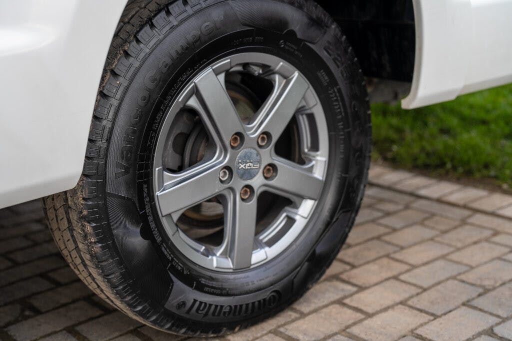 Close-up view of a silver alloy wheel on a white 2017 Swift Escape 664, fitted with a Continental VancoCamper tire. The vehicle is parked on a paved surface with a grassy area visible in the background.