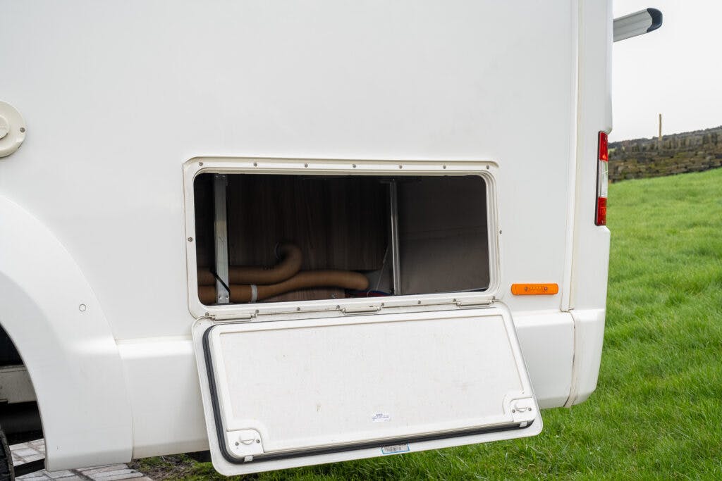 A white 2017 Swift Escape 664 motorhome with an open side compartment reveals storage space and plumbing. The compartment's door is hinged at the bottom and rests on the ground. The vehicle is parked on grass, with a part of a stone wall visible in the background.