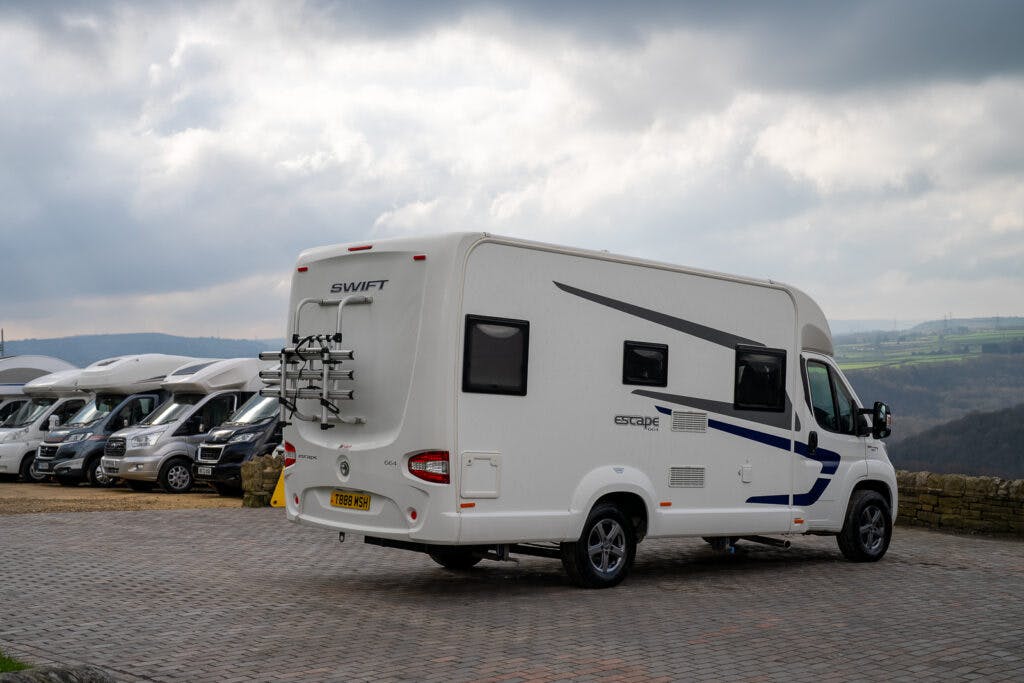 A white 2017 Swift Escape 664 motorhome is parked on a paved surface with scenic hills in the background. Several other motorhomes are parked in a row to the left. The sky is partly cloudy.