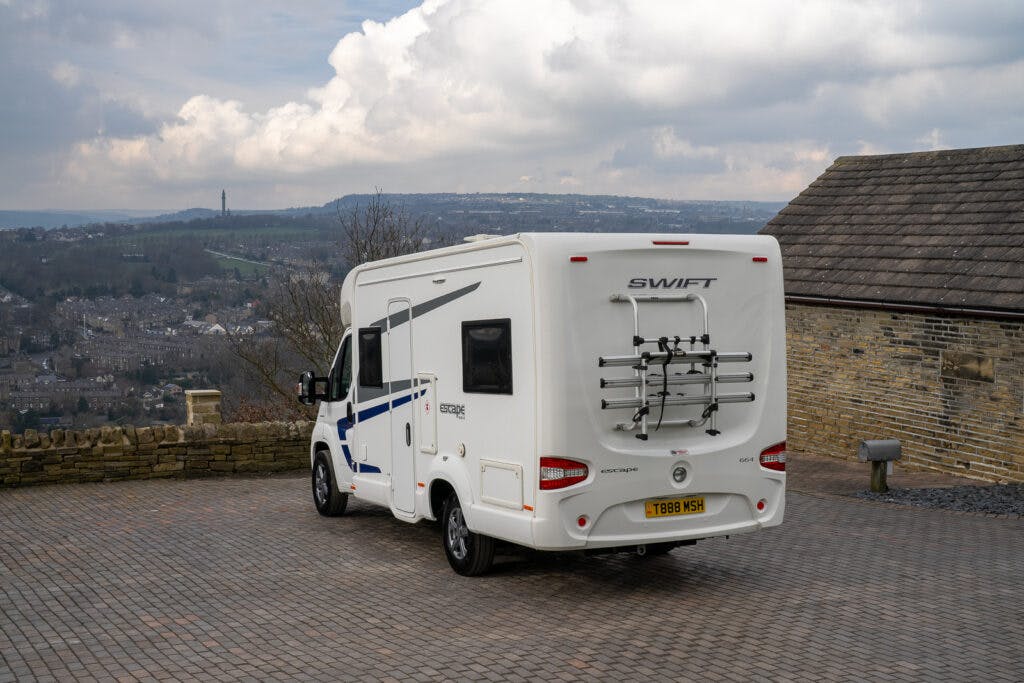 A white 2017 Swift Escape 664 motorhome is parked on a brick-paved area with a stone wall on the left and a building on the right. It has a bike rack and a visible license plate. The background shows a scenic view of a hilly landscape under a mostly cloudy sky.