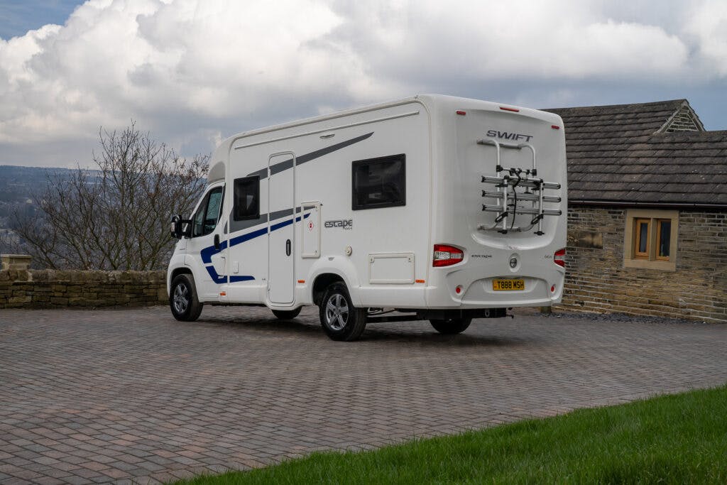A white 2017 Swift Escape 664 motorhome is parked on a stone-paved surface in front of a stone building. The vehicle has a bike rack on the rear and features a blue stripe along its side. The background includes a cloudy sky and a view of distant hills.