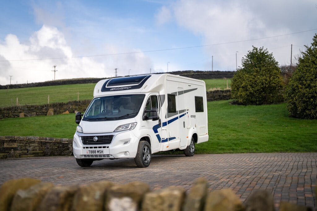 A 2017 Swift Escape 664 motorhome with a sleek black roof is parked on a cobblestone driveway. It features blue and black stripes on its side and a license plate reading "T 1888 NSH." The backdrop showcases a green grassy area, stone wall, small trees, and a partly cloudy sky.