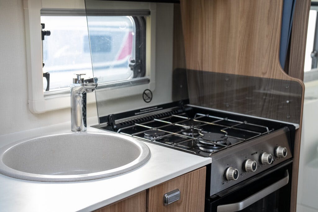 A compact kitchen inside the 2017 Swift Escape 664 mobile home features a round sink with a modern faucet, a gas stove with four burners, and an oven. The countertop is made of a light material, and there is a window above the sink. The cabinetry appears to be made of wood.