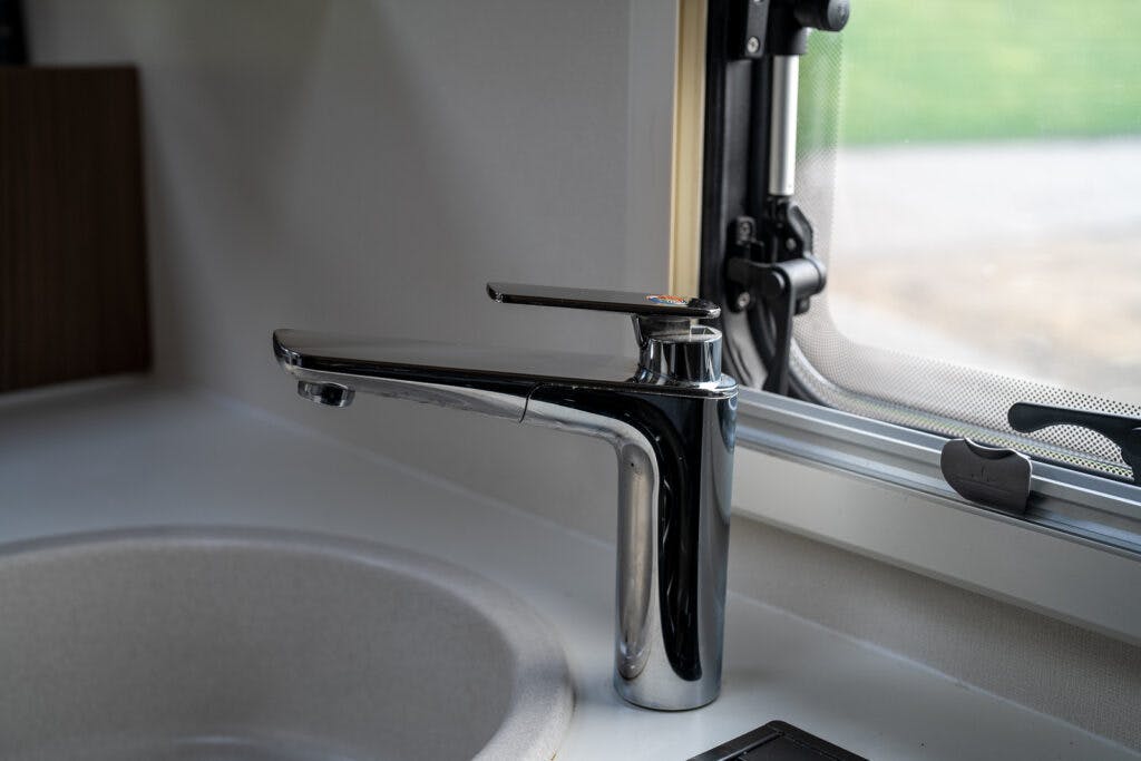 A close-up of a modern, chrome kitchen faucet mounted beside a round, beige sink in the 2017 Swift Escape 664. The faucet is positioned next to a window with a latch, allowing daylight to illuminate the area. The sink and faucet are part of a compact, sleek kitchen space.