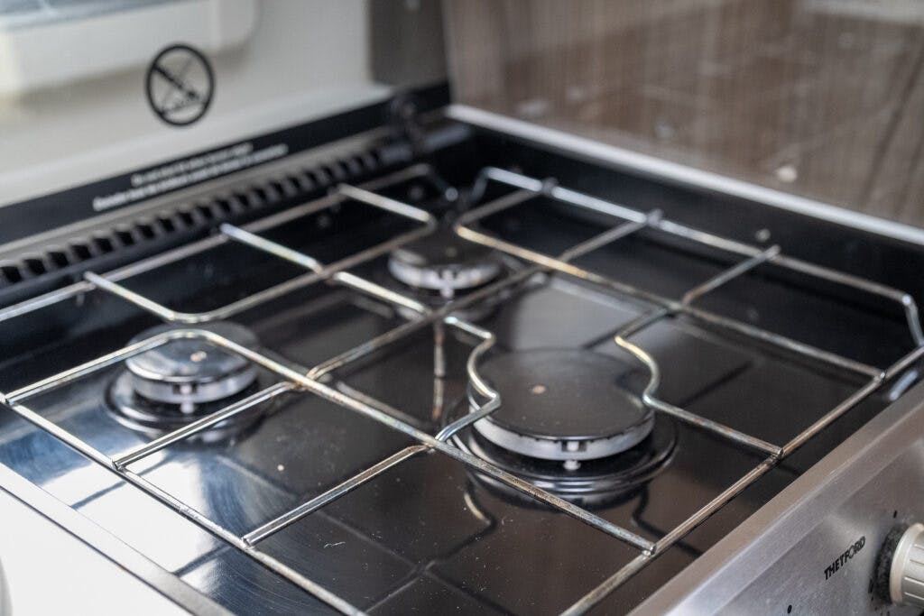 A close-up of a gas stove top with three burners in the 2017 Swift Escape 664. The stove has a metallic frame and black surface. The surrounding area is clean, and the burners appear to be in good condition. There is a "no smoking" sign on the wall behind the stove.
