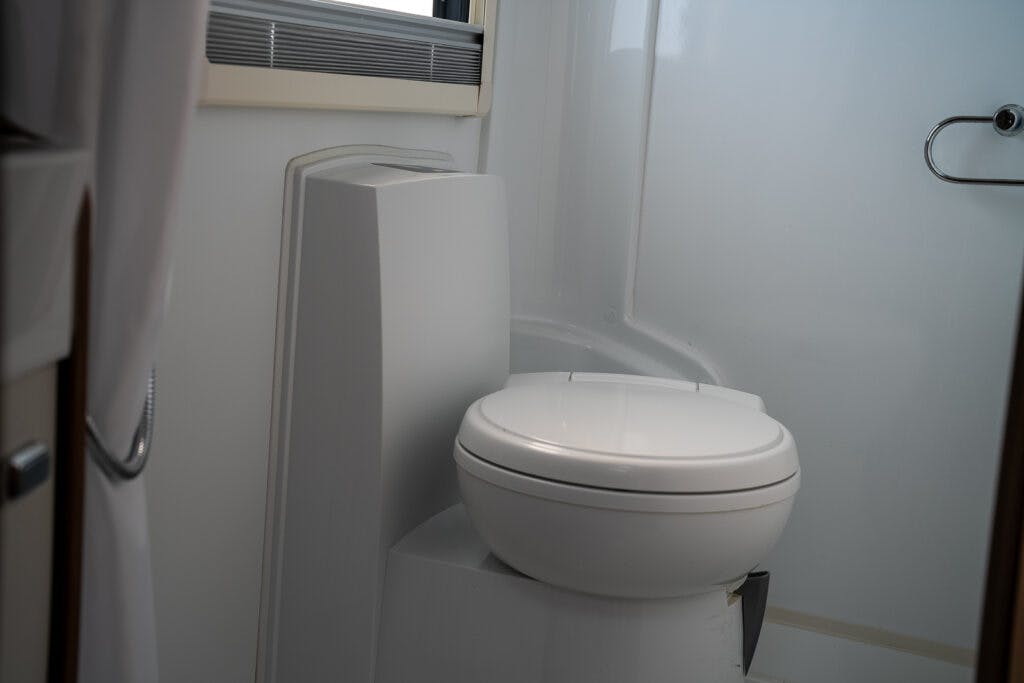 The image shows a compact bathroom inside the 2017 Swift Escape 664, featuring a white toilet situated within a white fiberglass shower stall. The toilet is mounted on an elevated platform, and a metal grab bar is affixed to the wall nearby. The bathroom has a clean, minimalist design.