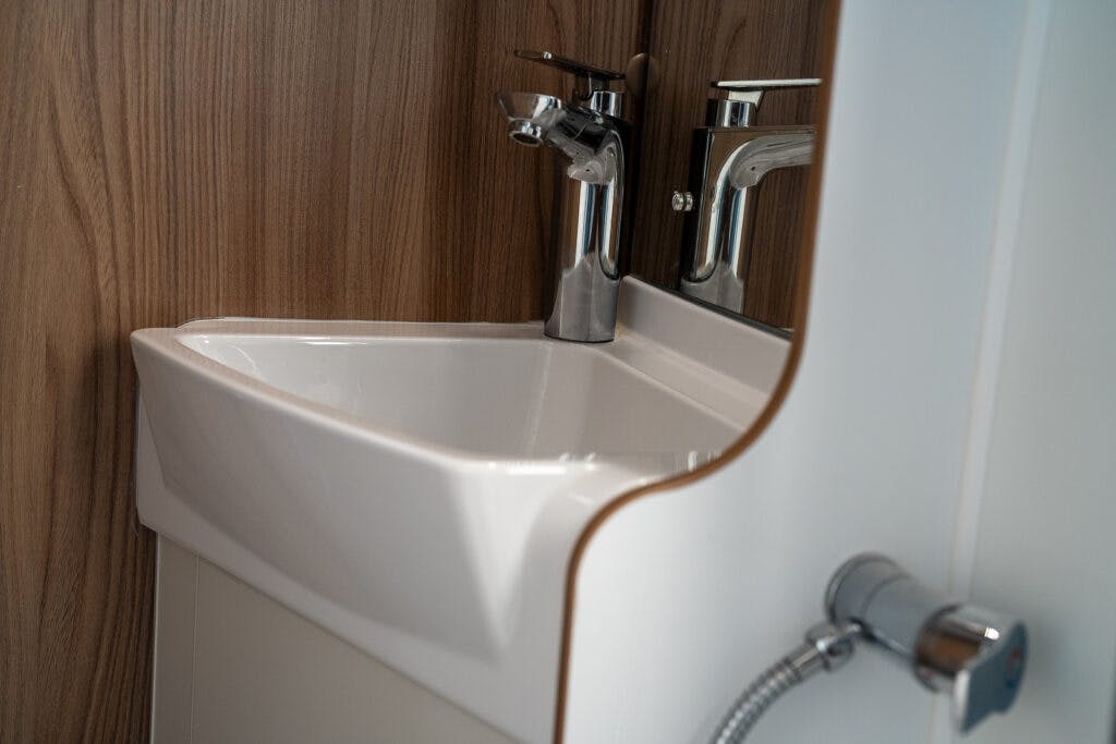 A close-up view of a modern white rectangular sink with a chrome faucet, reminiscent of the stylish design in the 2017 Swift Escape 664. The sink is mounted against a wooden wall panel with a partial white structure on the right. A metallic hose is visible in the bottom right corner.