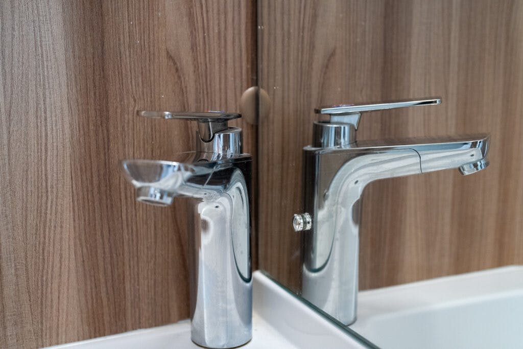 A modern chrome faucet with a lever handle is mounted on a bathroom sink inside the 2017 Swift Escape 664. It is reflected in a mirror, which is set against a wooden panel background. The reflection shows an identical faucet in the mirrored surface.