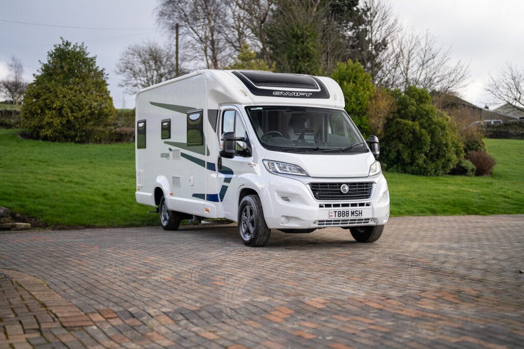 A white 2017 Swift Escape 664 motorhome is parked on a paved area with a grassy background. The motorhome has a panoramic front window and various graphics on the side. License plate reads "T688 WSH." Trees and bushes are visible in the background.