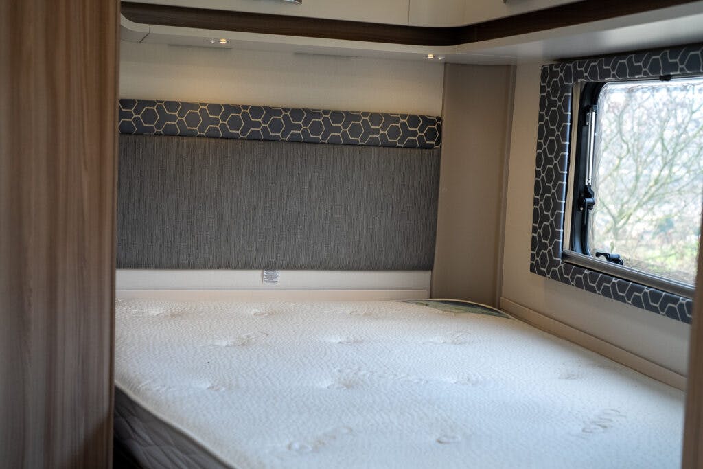 A neatly made bed with a white mattress in the 2017 Swift Escape 664 camper van. The interior features a padded headboard with a geometric pattern, a small window on the right side, and wood paneling on the left. The window provides a view of trees outside.