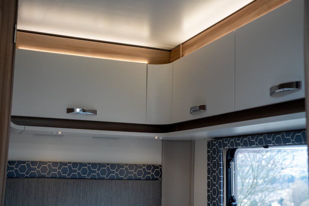 The image displays a modern interior view of a 2017 Swift Escape 664 RV. It shows a section of the ceiling with soft lighting, white overhead cabinets with metallic handles, and a window partially visible with a decorative geometric-patterned trim.