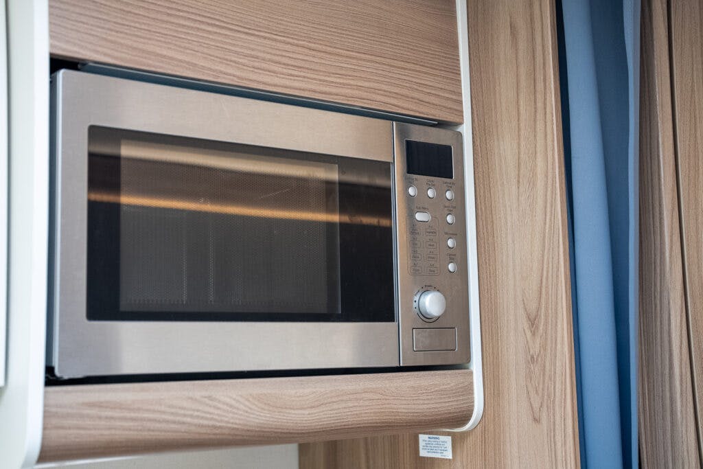 A silver microwave oven, reminiscent of the 2017 Swift Escape 664's sleek design, is built into a wooden kitchen cabinet. It features a digital display panel with various buttons and a glass door that allows the interior to be seen. A blue curtain is partially visible to the right of the microwave.