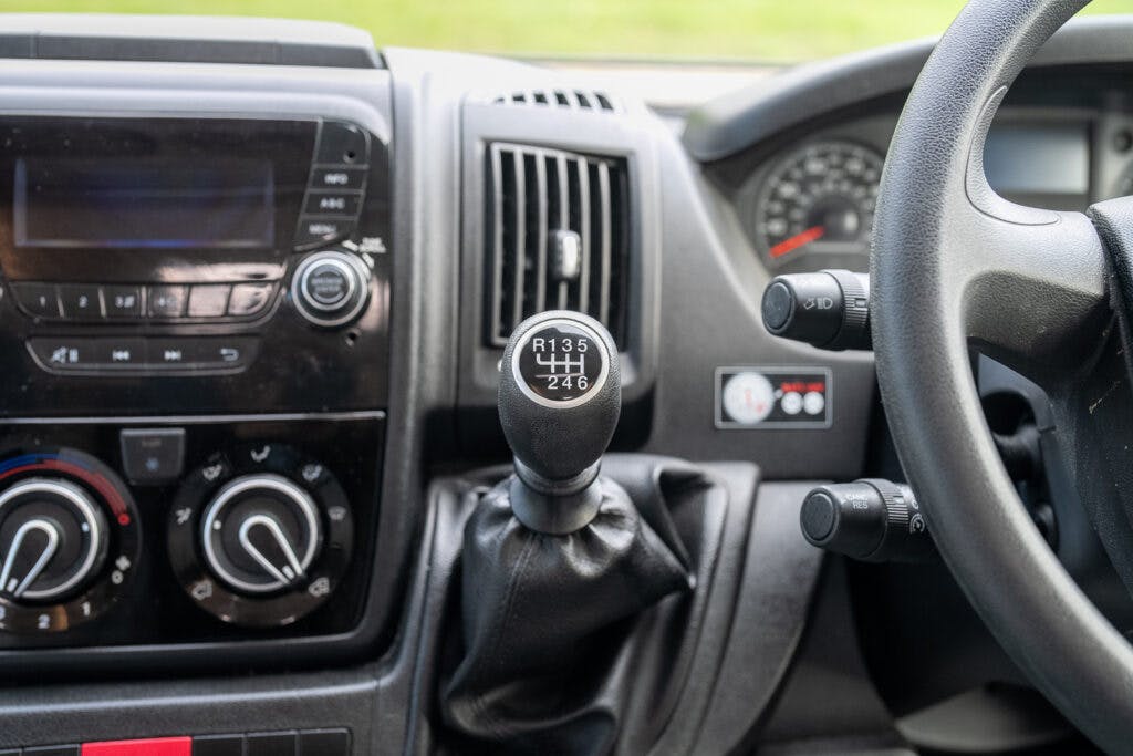 Close-up view of a 2017 Swift Escape 664’s interior showing the dashboard and gear shift. The gear shift includes an embossed pattern indicating seven possible gear positions including reverse. The dashboard features various controls, knobs, and a stereo system display.