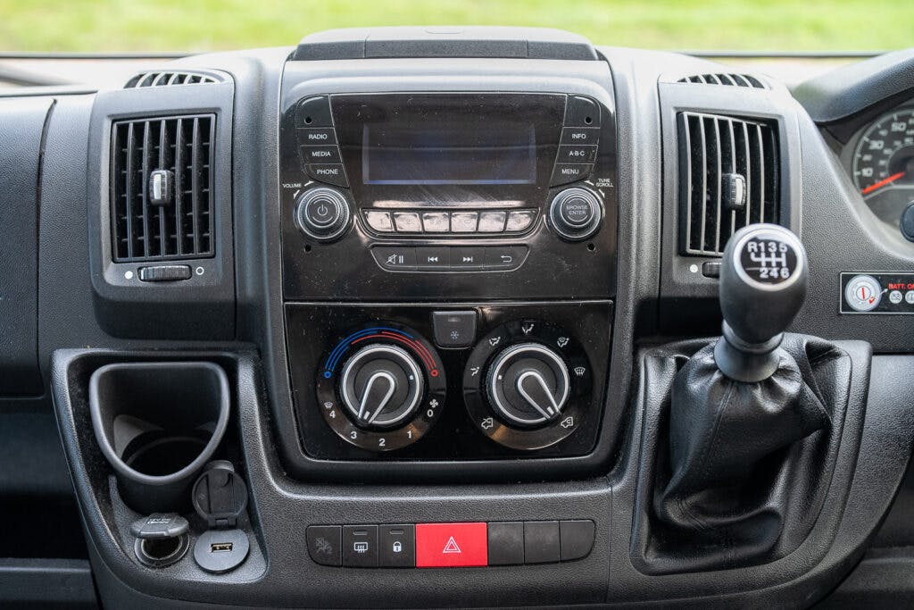A close-up view of the 2017 Swift Escape 664's center console. It features a digital display screen, two dials for air conditioning control, various buttons, a manual gear shift, and several power sockets. The hazard light button is prominently located at the bottom center.