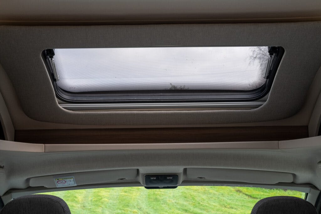 The image shows the interior roof of a 2017 Swift Escape 664 with an open rectangular sunroof. Beyond the sunroof, a cloudy sky and some tree branches are visible. The front headrests of the vehicle seats are partially seen at the bottom of the frame.