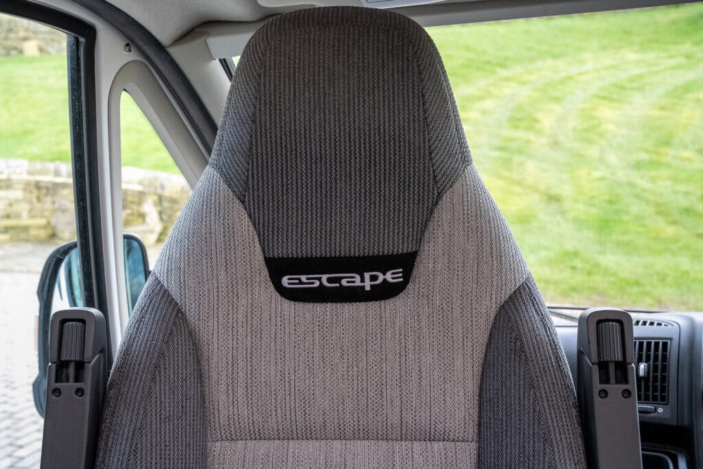 A close-up view of a car seat with grey upholstery. The seat has embroidery reading "Escape" on the backrest. In the background, through the window, green grass and part of a stone wall are visible—just one aspect of the stylish 2017 Swift Escape 664 interior.