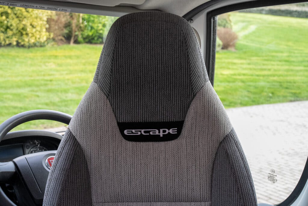 The image shows the driver's seat of a 2017 Swift Escape 664 from behind, highlighting its headrest which has "escape" embroidered on it. The interior view includes a portion of the steering wheel on the left. Through the window, a grassy area and pavement are visible outside.