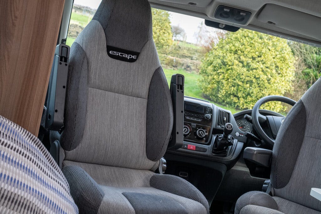 Interior view of a 2017 Swift Escape 664 camper van, featuring a grey driver's seat, a matching passenger seat labeled "escape," and a dashboard with various controls. The front windshield displays a green outdoor scene with trees and bushes.