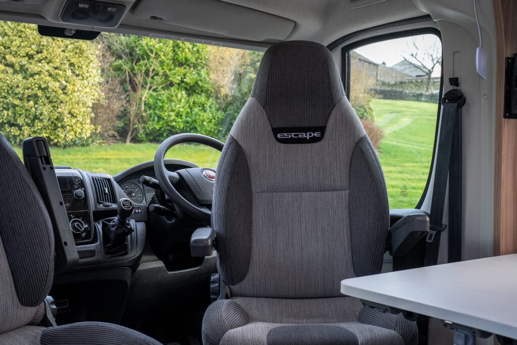 Interior view of a 2017 Swift Escape 664 motorhome featuring the driver's seat, steering wheel, and dashboard. The captain's chair labeled "escape" is covered in gray upholstery. Through the windows, a green lawn and some trees are visible outside.