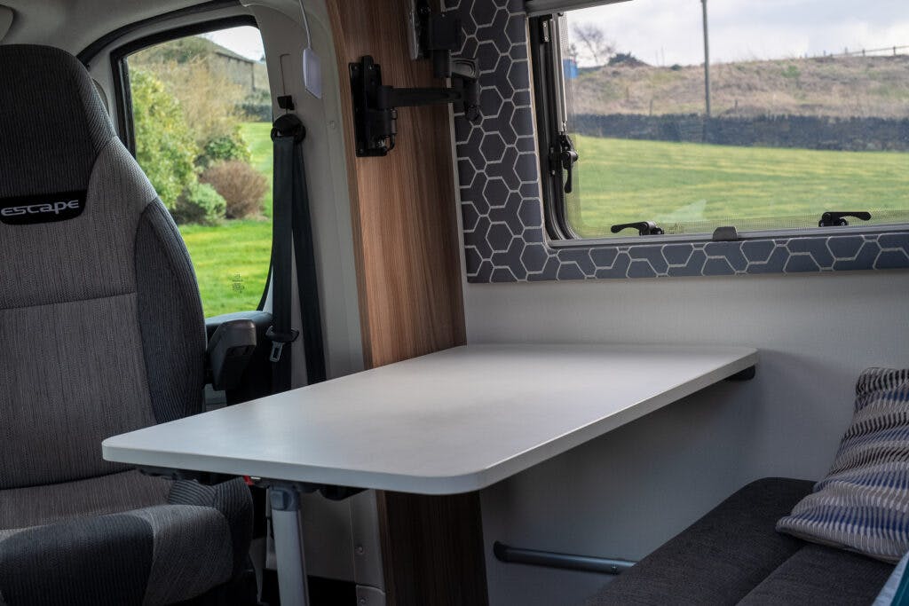 Interior view of a 2017 Swift Escape 664 camper van showing a table attached to the wall next to a window with a hexagonal-patterned shade. The table is positioned next to a gray passenger seat and a cushion is visible on a nearby bench. A grassy field is visible outside.