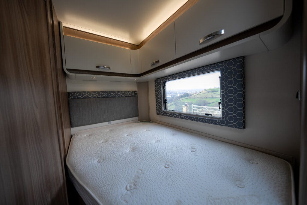 A single mattress on a wooden bed frame is situated in a compact bedroom with overhead storage cabinets. A large window with decorative trim provides a view of a hilly landscape outside. The room appears to be part of a 2017 Swift Escape 664 RV or mobile home.
