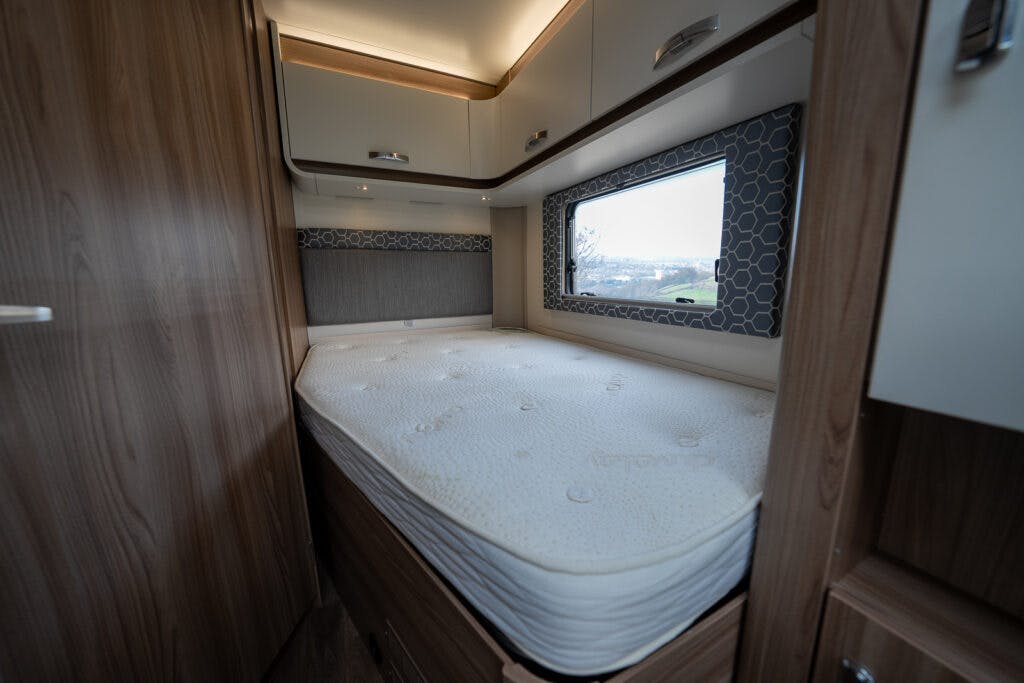 A small bedroom in the 2017 Swift Escape 664 features a neatly made bed with a white mattress. The walls are lined with wooden cabinets, and there's a window on one side with a geometric-patterned frame, allowing natural light into the interior.