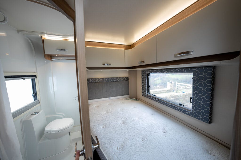 Image of a 2017 Swift Escape 664 RV interior featuring a bed with a white mattress and overhead storage cabinets. To the left, there is a small bathroom with a toilet visible. A window above the bed offers natural light with a view of the outside.