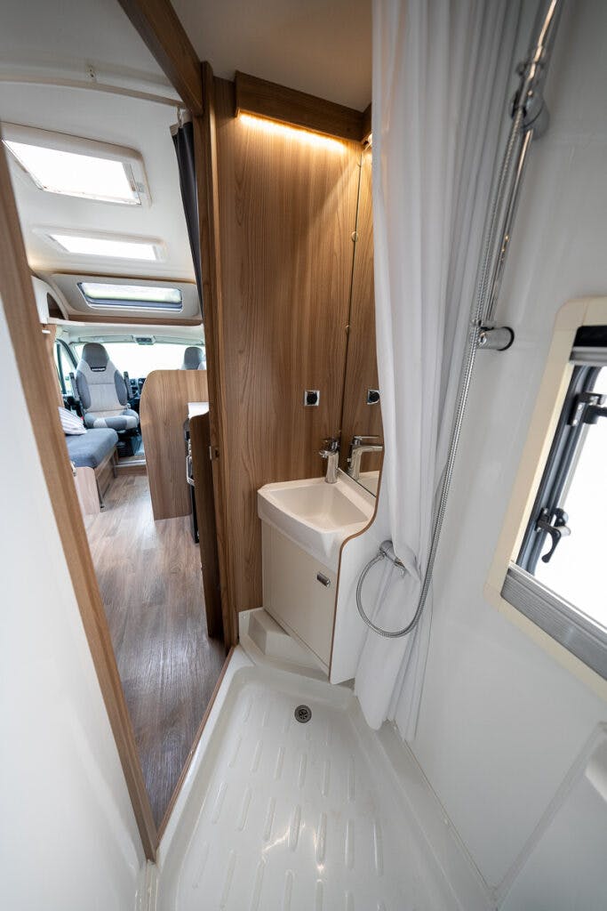 A view of the compact RV bathroom in the 2017 Swift Escape 664 features a small rectangular shower area with a white base and a curtain, adjacent to a tiny sink with a faucet and a small mirror above. The RV's main living area with seating is visible in the background.