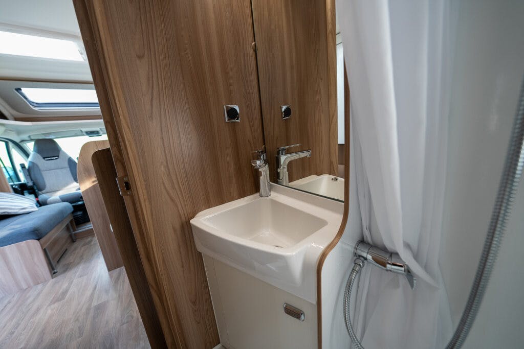 The image shows the interior of a 2017 Swift Escape 664 camper van, specifically the bathroom area with a sink and faucet. A wooden door and cabinetry are present, alongside a white curtain hinting at a shower space. The seating area in the van is partially visible in the background.