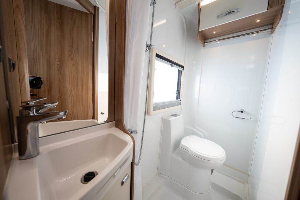 Modern RV bathroom in the 2017 Swift Escape 664 with a white toilet, a small sink with a chrome faucet, and a mirrored cabinet above. The right side wall has a frosted window, and a wooden door can be seen in the background. The space is compact and well-lit.
