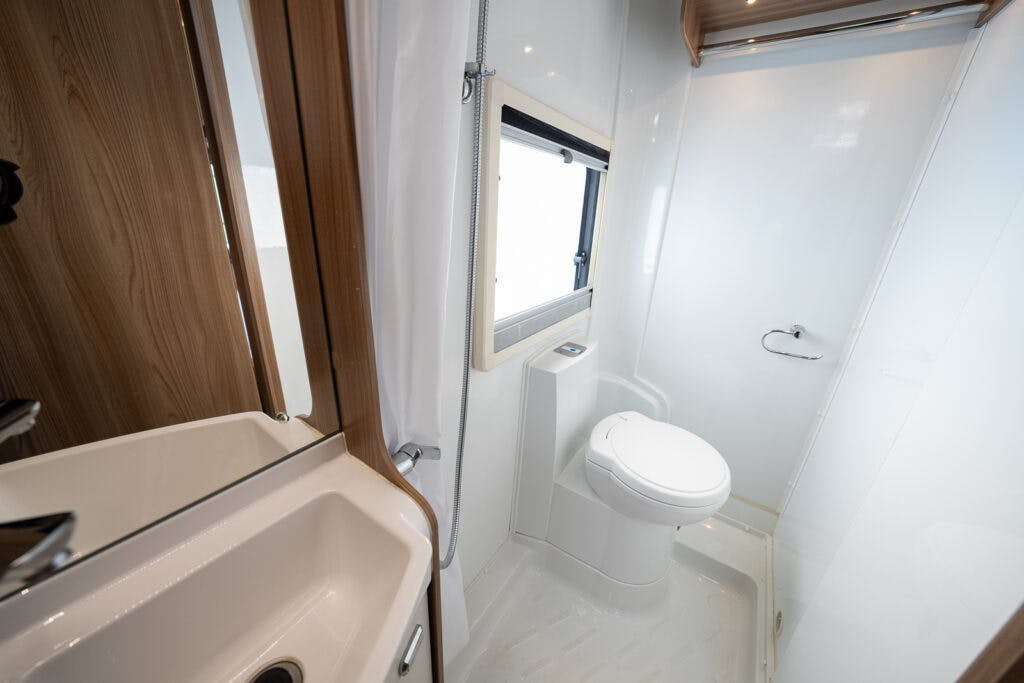 The 2017 Swift Escape 664 boasts a compact bathroom with wooden accents, featuring a white sink with a stainless steel faucet on the left, a window with a blind in the center, and a toilet with a closed lid on the right. The room has white walls and includes a small enclosed shower area.