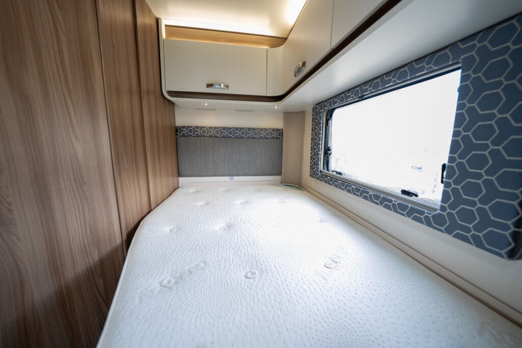The image showcases the interior of a 2017 Swift Escape 664 camper bedroom, featuring a single bed with a white mattress. Wooden paneling lines the left wall, and there are cabinets above the bed. A window with a patterned curtain graces the right side.