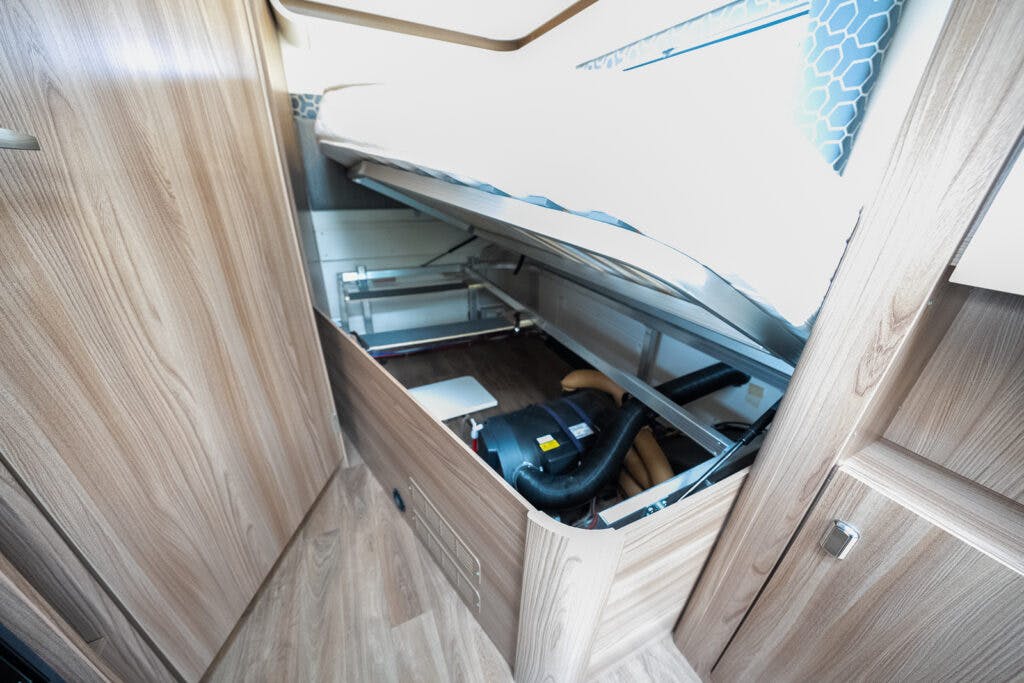 Interior of a 2017 Swift Escape 664 with a bed lifted to reveal storage space underneath. The storage area contains various items, including a black bag and some cables. The RV features light wood paneling and cabinetry.
