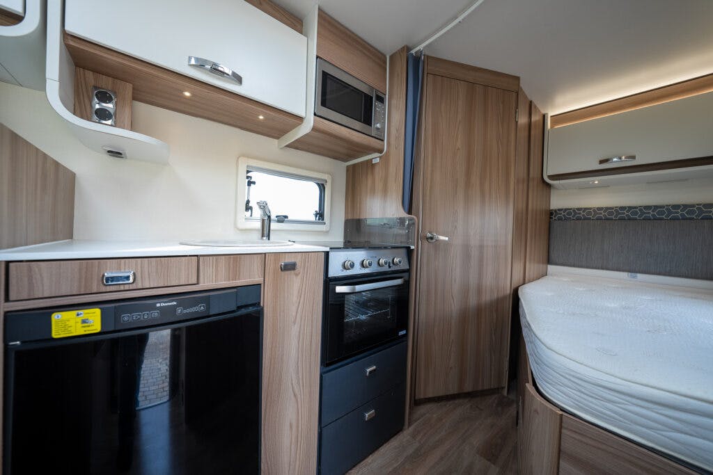 Interior of the 2017 Swift Escape 664 camper van featuring a small kitchen with a sink, stovetop, oven, microwave, and storage cabinets. Adjacent is a compact sleeping area with a mattress. The decor includes light wood finishes and minimalistic design elements.