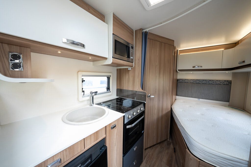 The image shows the interior of a 2017 Swift Escape 664 camper van featuring a kitchenette with a sink, stovetop, and microwave, alongside a small oven. Adjacent to the kitchenette is a cozy sleeping area with a bed and cabinets for storage. The decor is light wood and white.
