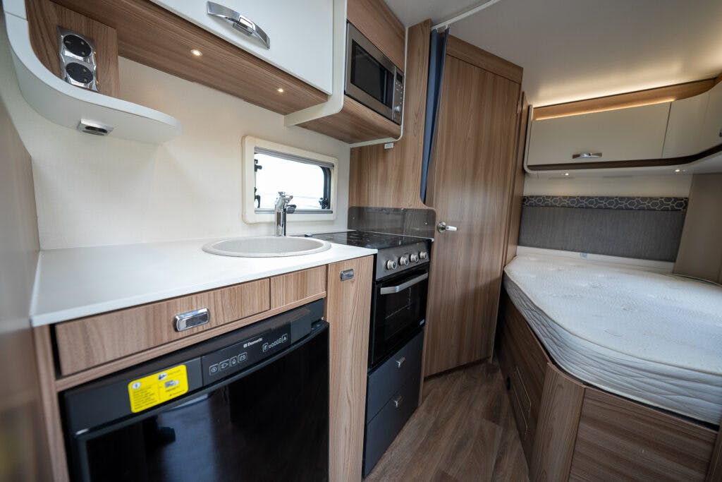 The image shows the interior of a 2017 Swift Escape 664 camper van featuring a compact kitchenette with a sink, stove, oven, and microwave. Adjacent to the kitchenette is a small bed. The cabinetry is wood-themed, and there is a window above the sink.