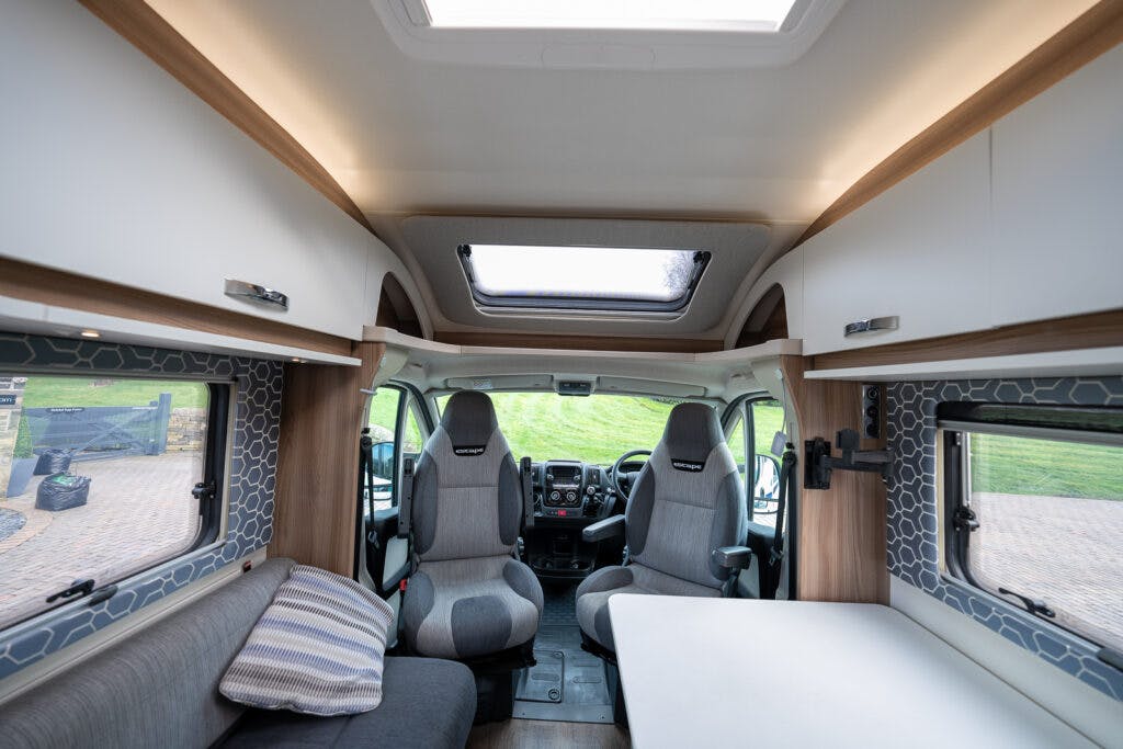 Interior view of the 2017 Swift Escape 664 camper van showing two front seats, a small table, a sofa with a striped cushion to the left, and overhead storage cabinets. A large skylight is visible in the ceiling, and the windows provide views of greenery and a cobblestone path outside.