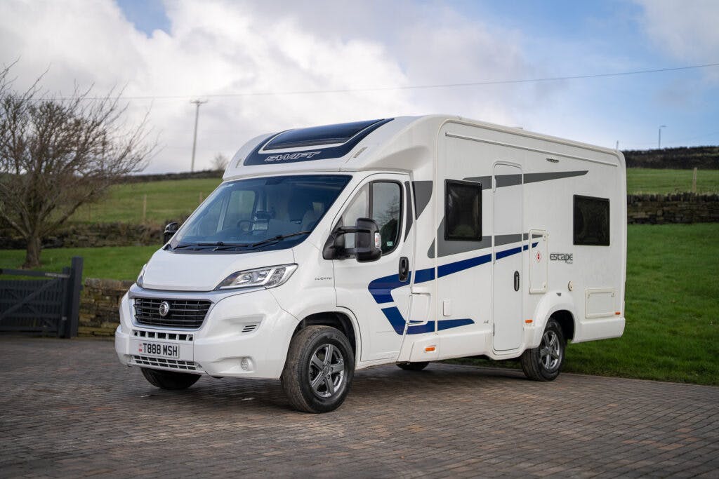 A 2017 Swift Escape 664 motorhome in white and blue is parked on a paved area. The vehicle, sporting the registration plate "T1888 MSH," features various windows and storage compartments. In the background, a grassy field stretches under a cloudy sky.