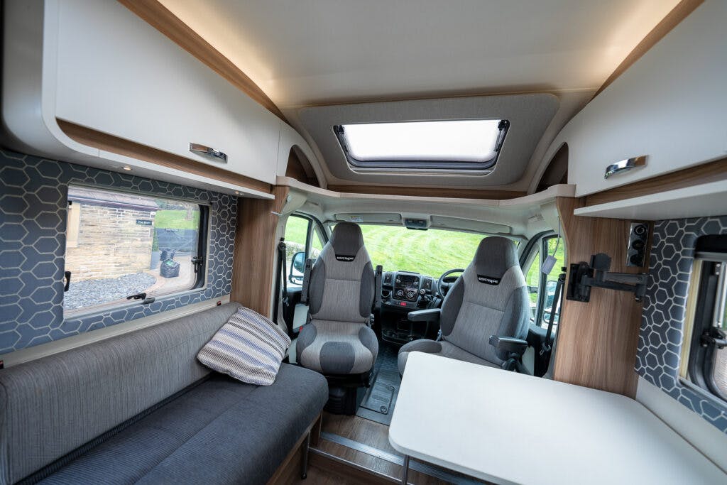 Interior of the 2017 Swift Escape 664 featuring two captain chairs, a grey sofa with a striped pillow, and a small table. The walls have a hexagonal pattern, and there is a skylight on the ceiling. The windows offer a view of the grassy outdoors.