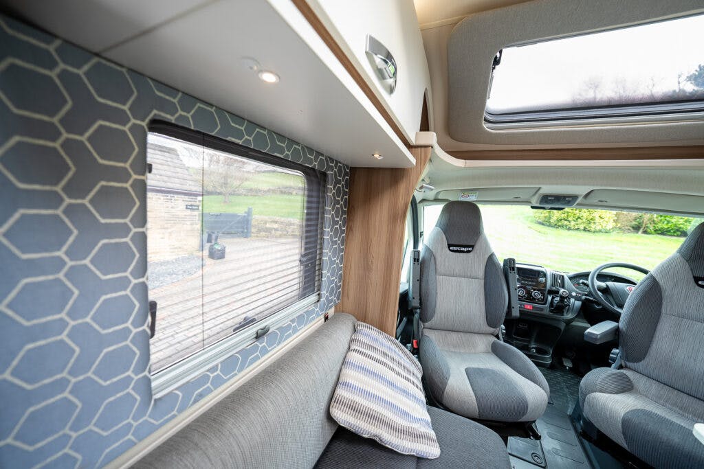 The interior of the 2017 Swift Escape 664 motorhome showcases a driver's seat, passenger seat, and a seating area with a gray couch and striped cushion. The interior walls feature a blue hexagonal pattern, complemented by a sunroof and window offering outside views.