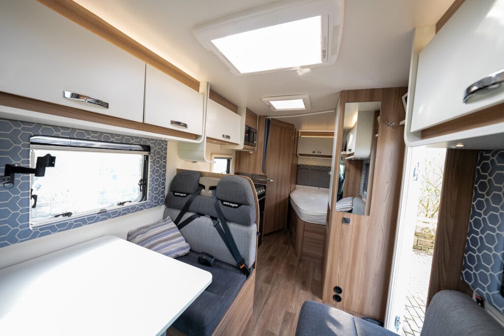 The image shows the interior of a 2017 Swift Escape 664 camper van. It has wood-paneled walls, a small dining area with bench seating, overhead storage cabinets, and a sleeping area with a bed. There's a skylight on the ceiling and a window on the side.