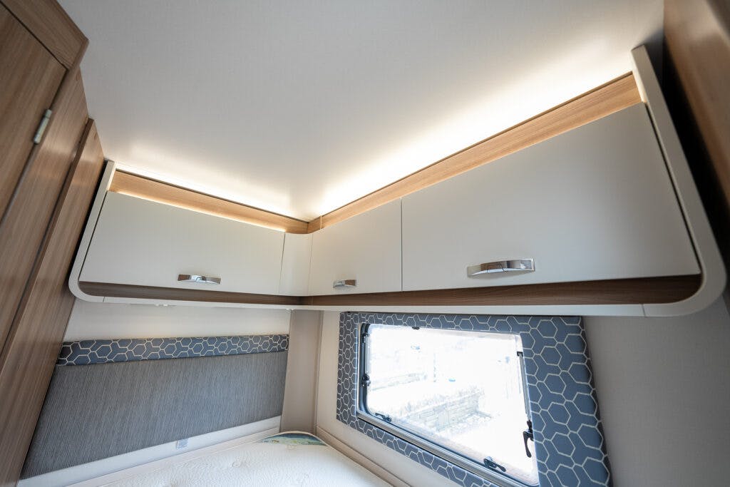 Interior of a 2017 Swift Escape 664 camper van showing upper storage cabinets with a wooden finish and under-cabinet lighting. A bed is partially visible below with a patterned headboard and a window to the right. The walls and ceiling are light-colored.