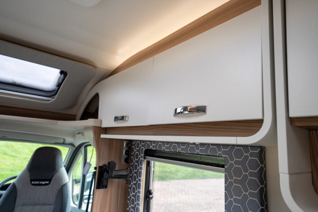 The image shows the interior of a 2017 Swift Escape 664 camper van, focusing on the top cabinets above a window. The cabinets are white with wooden trim and silver handles. Below, a patterned backsplash is visible. The driver's seat, marked as "Escape," is partially seen on the left.