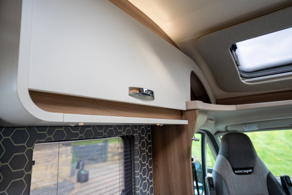 Interior shot of a 2017 Swift Escape 664 campervan featuring a storage cabinet with a modern design. The cabinet has wooden accents and a sleek metallic handle. There is a window with a hexagon-patterned curtain, and an overhead skylight is partially visible.